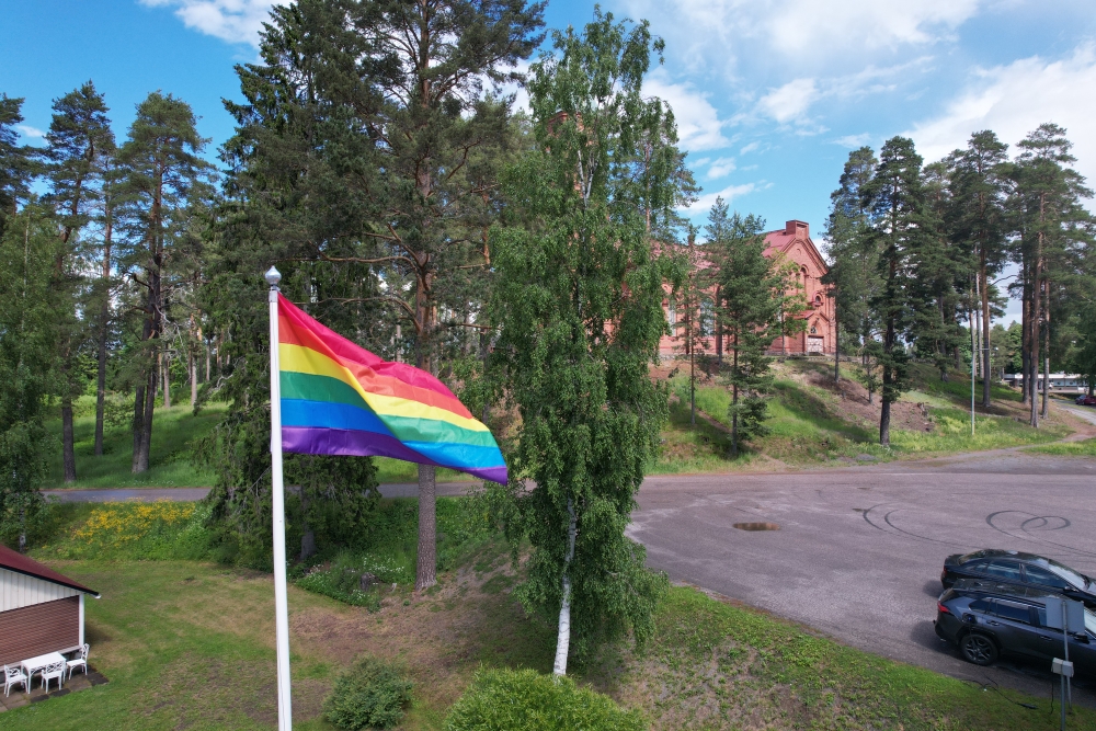 Sateenkaarilippu. Taustalla lopen kirkon parkkipaikkaa ja kirkko puiden takana